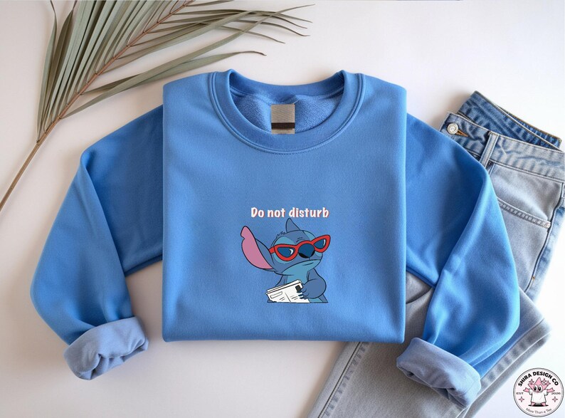 Peut inclure: Un sweat-shirt bleu clair avec un personnage de dessin anim&eacute; portant des lunettes de soleil rouges et tenant un papier, avec le texte "Do not disturb". Le sweat-shirt est pli&eacute; &agrave; c&ocirc;t&eacute; d'un jean bleu.