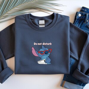 Peut inclure: Un sweat-shirt bleu marine avec un personnage de dessin anim&eacute; Stitch portant des lunettes de soleil rouges et tenant un journal. Le texte "Do not disturb" est au-dessus du personnage. Le sweat-shirt est associ&eacute; &agrave; un jean bleu.