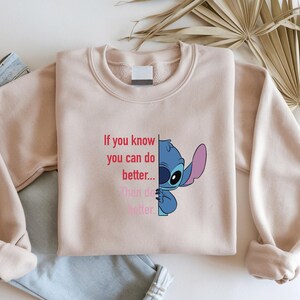 May include: A light pink sweatshirt featuring a blue cartoon character and the text "If you know you can do better... Then do better." The sweatshirt is folded on top of a pair of blue jeans.
