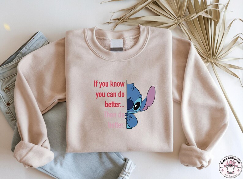 May include: A light pink sweatshirt featuring a blue cartoon character and the text "If you know you can do better... Then do better." The sweatshirt is folded on top of a pair of blue jeans.