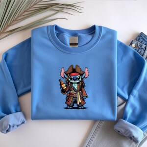 May include: A light blue sweatshirt featuring a cartoon character dressed as a pirate, holding a bottle. The character has blue fur, a brown pirate hat, and a red bandana. The sweatshirt is folded with a pair of jeans.