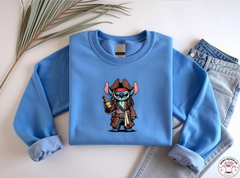 May include: A light blue sweatshirt featuring a cartoon character dressed as a pirate, holding a bottle. The character has blue fur, a brown pirate hat, and a red bandana. The sweatshirt is folded with a pair of jeans.