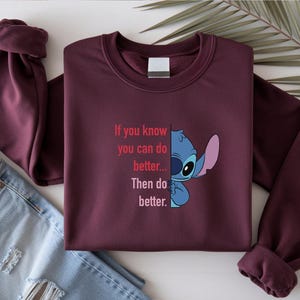 May include: Burgundy sweatshirt featuring a graphic of Stitch from Lilo & Stitch and the text "If you know you can do better... Then do better." The long-sleeved top is folded with a pair of blue jeans.