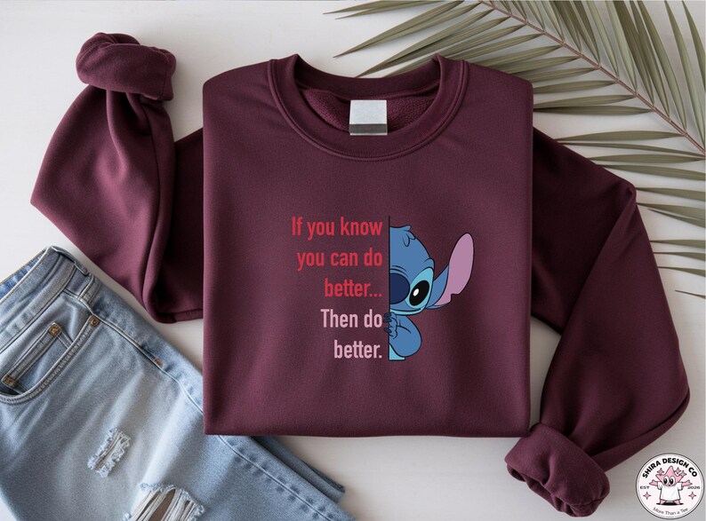 May include: Burgundy sweatshirt featuring a graphic of Stitch from Lilo & Stitch and the text "If you know you can do better... Then do better." The long-sleeved top is folded with a pair of blue jeans.