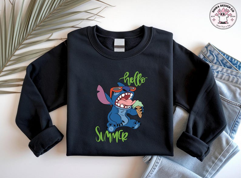 May include: Black crew neck sweatshirt with a cartoon character wearing sunglasses and holding an ice cream cone. The words "hello" and "summer" are written in green script. The sweatshirt is paired with blue jeans.