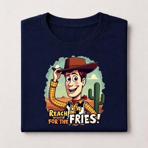 Op de afbeelding: Marineblauw T-shirt met een cartoon illustratie van een cowboy met een bruine hoed, geel geruit overhemd en rode halsdoek. De tekst "REACH FOR THE FRIES!" staat eronder.