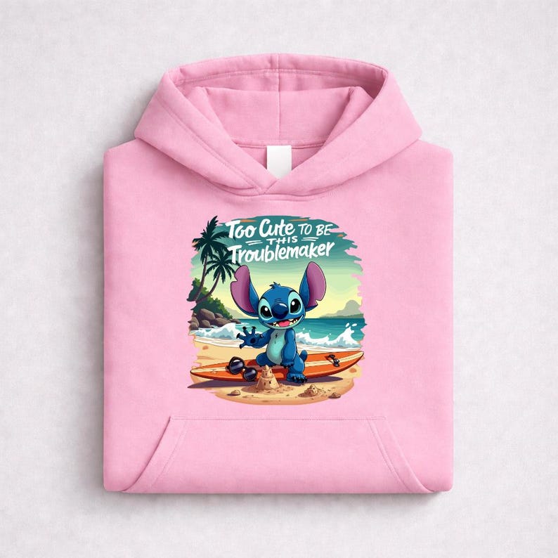 Op de afbeelding: Een lichtroze hoodie met een afbeelding van een blauw stripfiguur op een surfplank op het strand. Het personage lacht, met de tekst "Too Cute to be this Troublemaker" erboven. De hoodie heeft een voorzak en een capuchon met trekkoord.