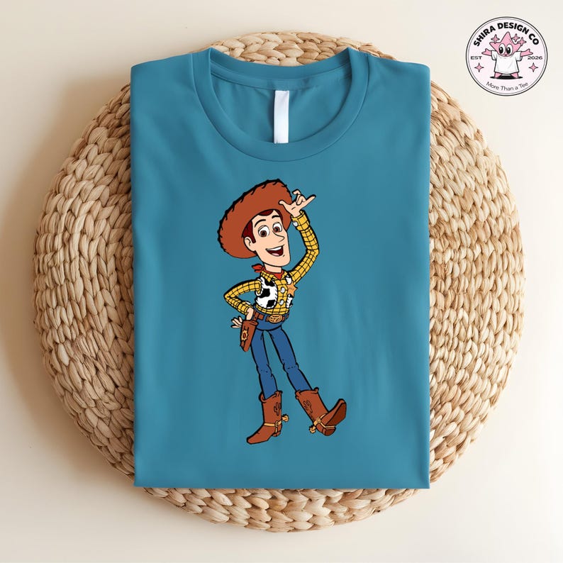 May include: Teal blue t-shirt featuring a cartoon illustration of Woody from Toy Story. Woody is wearing a brown cowboy hat, a yellow plaid shirt, blue jeans, and brown boots. The shirt has the text "Shira Design Co" and "More Than a Tee" on the upper right.