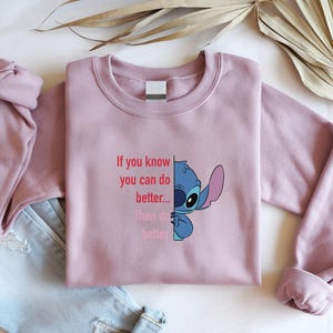 May include: A mauve crewneck sweatshirt featuring a blue cartoon character and the text "If you know you can do better... Then do better". The design is on the front of the sweatshirt. A pair of blue jeans is visible in the background.