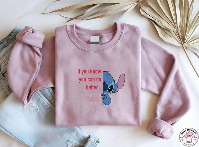 May include: A mauve crewneck sweatshirt featuring a blue cartoon character and the text "If you know you can do better... Then do better". The design is on the front of the sweatshirt. A pair of blue jeans is visible in the background.