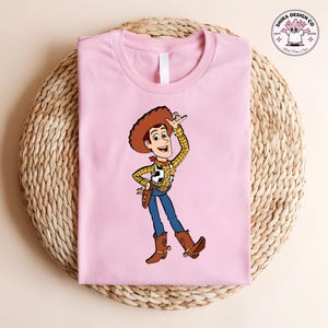 May include: A light pink t-shirt featuring a cartoon illustration of Woody from Toy Story. Woody is wearing a brown cowboy hat, a yellow plaid shirt, blue jeans, and brown boots. The shirt is folded on a woven surface.