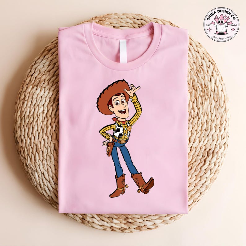 May include: A light pink t-shirt featuring a cartoon illustration of Woody from Toy Story. Woody is wearing a brown cowboy hat, a yellow plaid shirt, blue jeans, and brown boots. The shirt is folded on a woven surface.