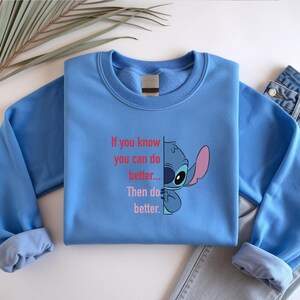 May include: A light blue sweatshirt featuring a graphic of Stitch from Lilo & Stitch, with the text "If you know you can do better... Then do better." The design includes Stitch's face and ear. A pair of blue jeans is visible.