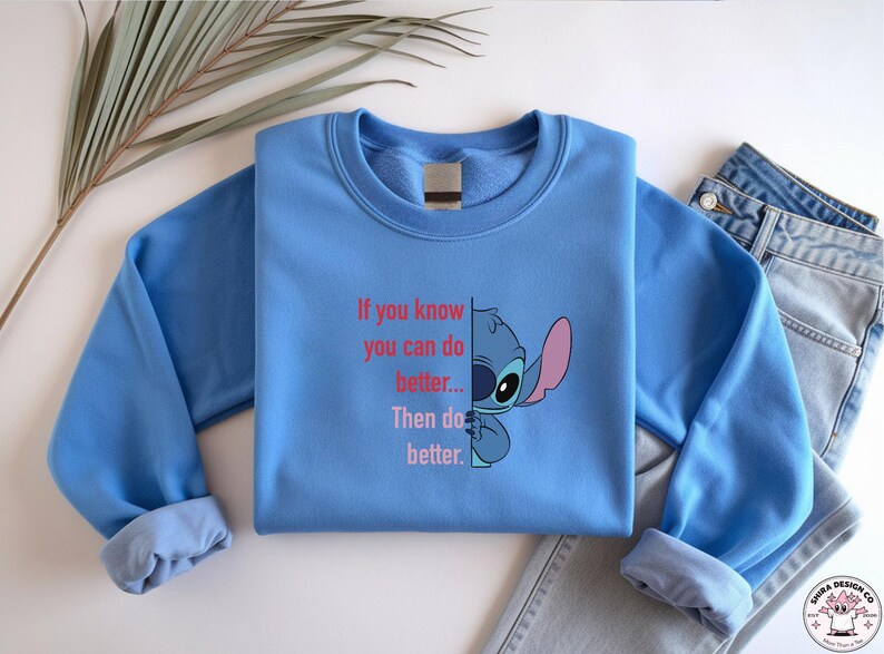 May include: A light blue sweatshirt featuring a graphic of Stitch from Lilo & Stitch, with the text "If you know you can do better... Then do better." The design includes Stitch's face and ear. A pair of blue jeans is visible.
