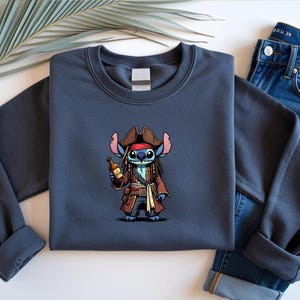 May include: A navy blue sweatshirt featuring a cartoon character dressed as a pirate, holding a bottle. The character has blue skin, large ears, and is wearing a brown pirate hat and coat. The sweatshirt is paired with blue jeans.