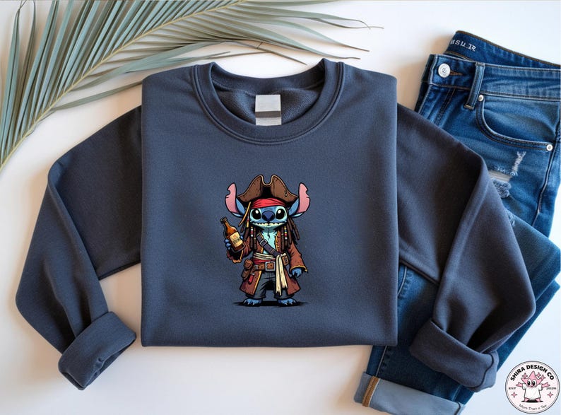 May include: A navy blue sweatshirt featuring a cartoon character dressed as a pirate, holding a bottle. The character has blue skin, large ears, and is wearing a brown pirate hat and coat. The sweatshirt is paired with blue jeans.