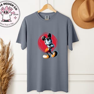 May include: A grey t-shirt with a cartoon Mickey Mouse design, posed dynamically against a red circle. The shirt has a crew neck and short sleeves. The image also includes a logo that says "Shira Design Co" and "More Than a Tee".