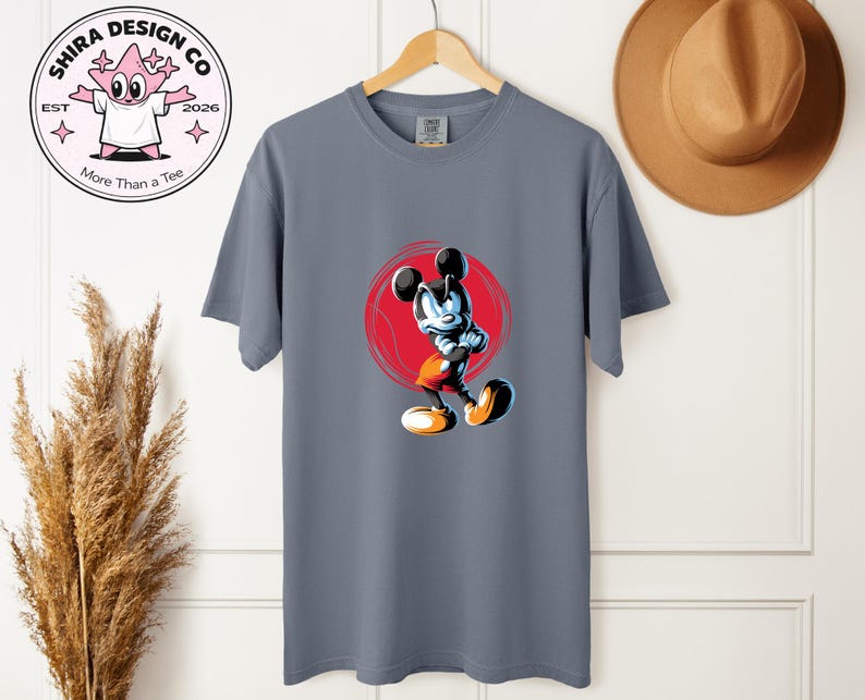 May include: A grey t-shirt with a cartoon Mickey Mouse design, posed dynamically against a red circle. The shirt has a crew neck and short sleeves. The image also includes a logo that says "Shira Design Co" and "More Than a Tee".