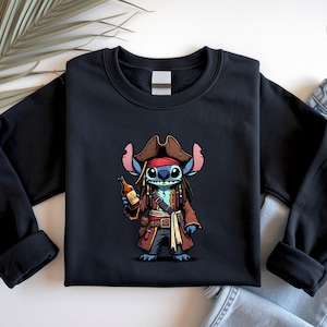 May include: Black sweatshirt featuring a cartoon character dressed as a pirate, holding a bottle. The character is blue with large ears and dreadlocks, wearing a brown pirate hat and coat. The design is centered on the front of the sweatshirt.