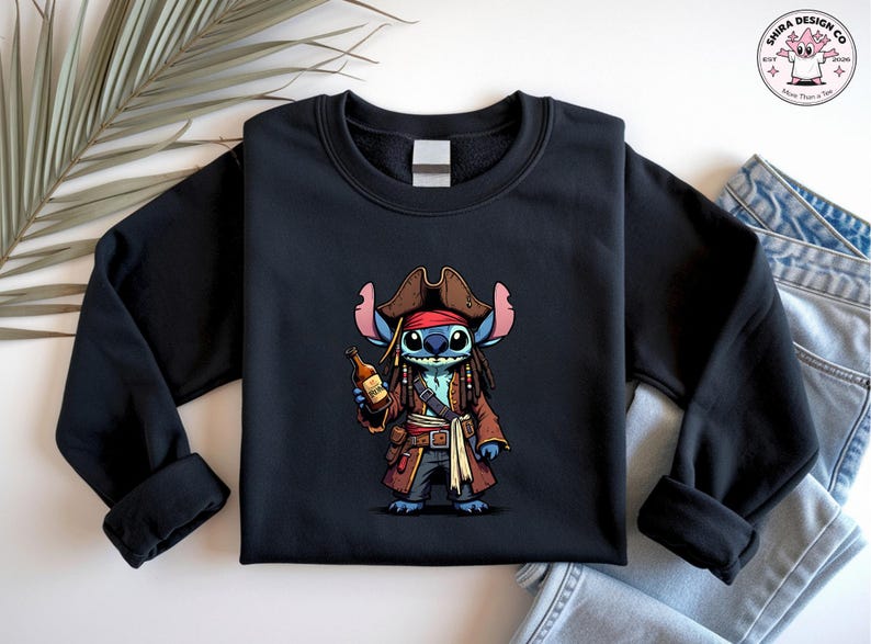 May include: Black sweatshirt featuring a cartoon character dressed as a pirate, holding a bottle. The character is blue with large ears and dreadlocks, wearing a brown pirate hat and coat. The design is centered on the front of the sweatshirt.