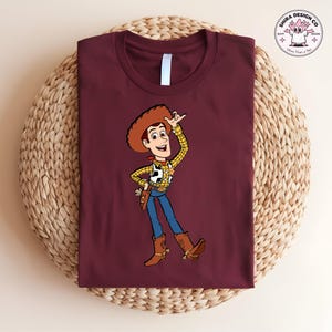 May include: Burgundy t-shirt featuring a cartoon illustration of a cowboy character. The cowboy is wearing a brown hat, a yellow plaid shirt, blue jeans, and brown boots. The shirt has a white collar and the image is on a woven surface.
