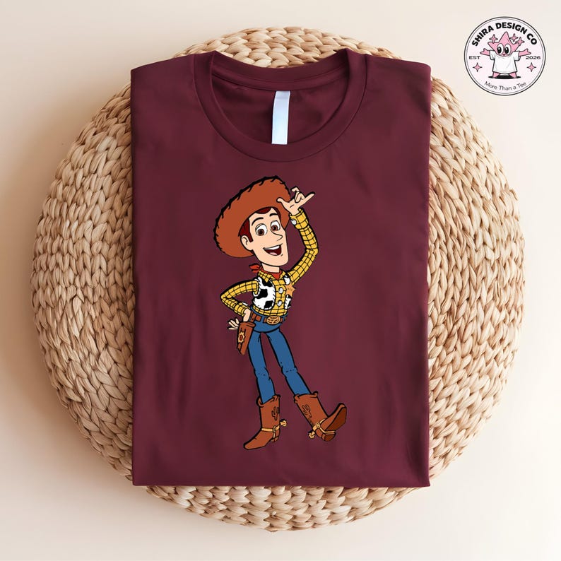May include: Burgundy t-shirt featuring a cartoon illustration of a cowboy character. The cowboy is wearing a brown hat, a yellow plaid shirt, blue jeans, and brown boots. The shirt has a white collar and the image is on a woven surface.