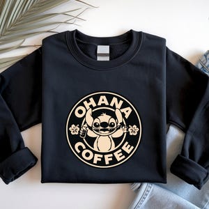 May include: Black sweatshirt featuring a circular design with the words "OHANA COFFEE" around a cartoon character holding a coffee cup and flowers. The design is in a cream color. The sweatshirt has long sleeves.
