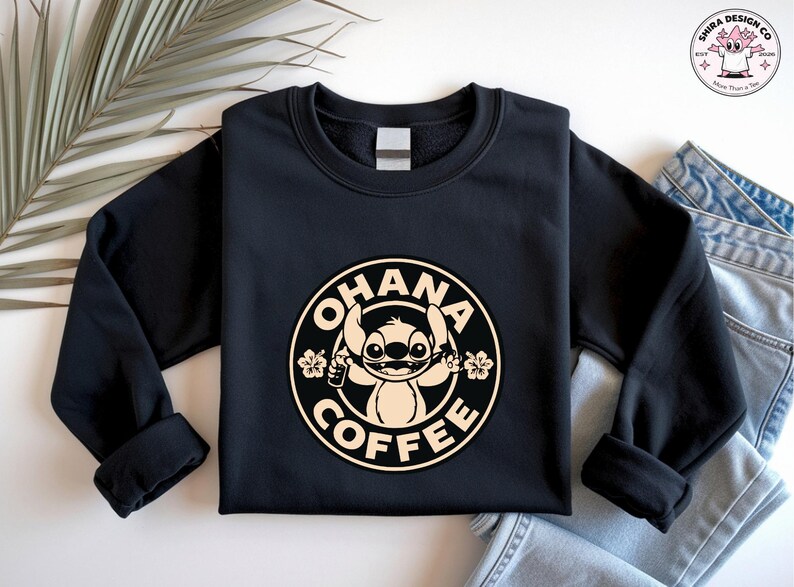 May include: Black sweatshirt featuring a circular design with the words "OHANA COFFEE" around a cartoon character holding a coffee cup and flowers. The design is in a cream color. The sweatshirt has long sleeves.