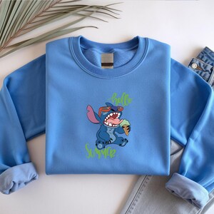May include: A pale blue sweatshirt with a cartoon character wearing sunglasses and holding an ice cream. The words "hello" and "summer" are written in green. The sweatshirt is folded on a white surface next to a pair of blue jeans.