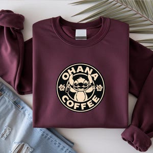 May include: A maroon sweatshirt with a circular graphic featuring the character Stitch from Lilo & Stitch, with the words "OHANA COFFEE". The graphic is in black and beige. The sweatshirt is folded next to a pair of blue jeans.