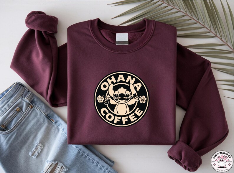 May include: A maroon sweatshirt with a circular graphic featuring the character Stitch from Lilo & Stitch, with the words "OHANA COFFEE". The graphic is in black and beige. The sweatshirt is folded next to a pair of blue jeans.