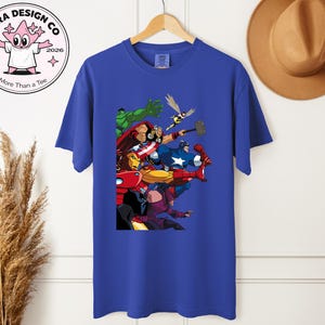 Może przedstawiać: Kr&oacute;lewski niebieski t-shirt z kolorową grafiką superbohater&oacute;w, w tym Hulka, Iron Mana i Kapitan Ameryki. Koszulka wisi na drewnianym wieszaku. Logo marki jest widoczne w lewym g&oacute;rnym rogu. Koszulka jest w żywym odcieniu niebieskiego.