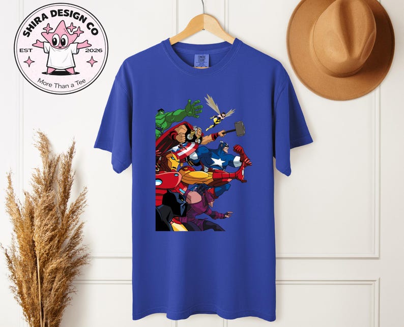 Może przedstawiać: Kr&oacute;lewski niebieski t-shirt z kolorową grafiką superbohater&oacute;w, w tym Hulka, Iron Mana i Kapitan Ameryki. Koszulka wisi na drewnianym wieszaku. Logo marki jest widoczne w lewym g&oacute;rnym rogu. Koszulka jest w żywym odcieniu niebieskiego.