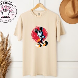 May include: Beige t-shirt with a cartoon Mickey Mouse design inside a red circle. The graphic depicts Mickey in a dynamic pose with black, blue, orange, and white colours. The shirt hangs on a wooden hanger. The brand logo is visible in the upper left corner.