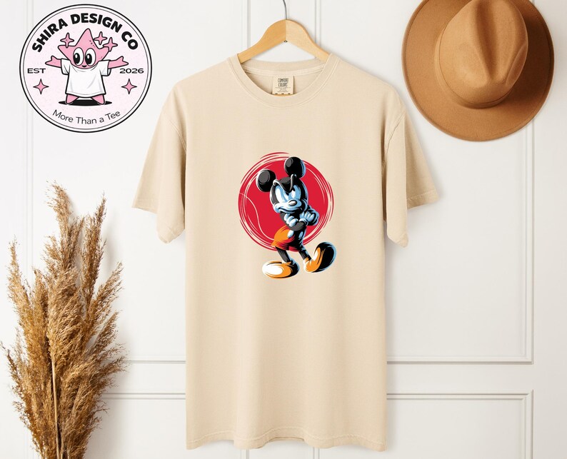 May include: Beige t-shirt with a cartoon Mickey Mouse design inside a red circle. The graphic depicts Mickey in a dynamic pose with black, blue, orange, and white colours. The shirt hangs on a wooden hanger. The brand logo is visible in the upper left corner.
