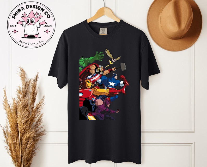 Może przedstawiać: Czarny t-shirt z kolorową grafiką superbohater&oacute;w, w tym Kapitanem Ameryką i Iron Manem. Projekt jest wyświetlany na drewnianym wieszaku. Logo marki jest widoczne w lewym g&oacute;rnym rogu. Koszulka jest częścią kolekcji odzieżowej.