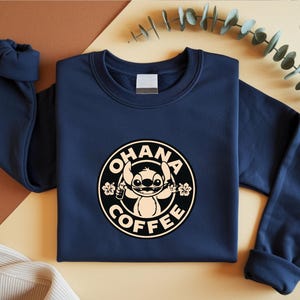 May include: Navy blue sweatshirt featuring a circular graphic with the text "OHANA COFFEE" and a cartoon character. The design is in black and beige. The sweatshirt has long sleeves and a crew neck.
