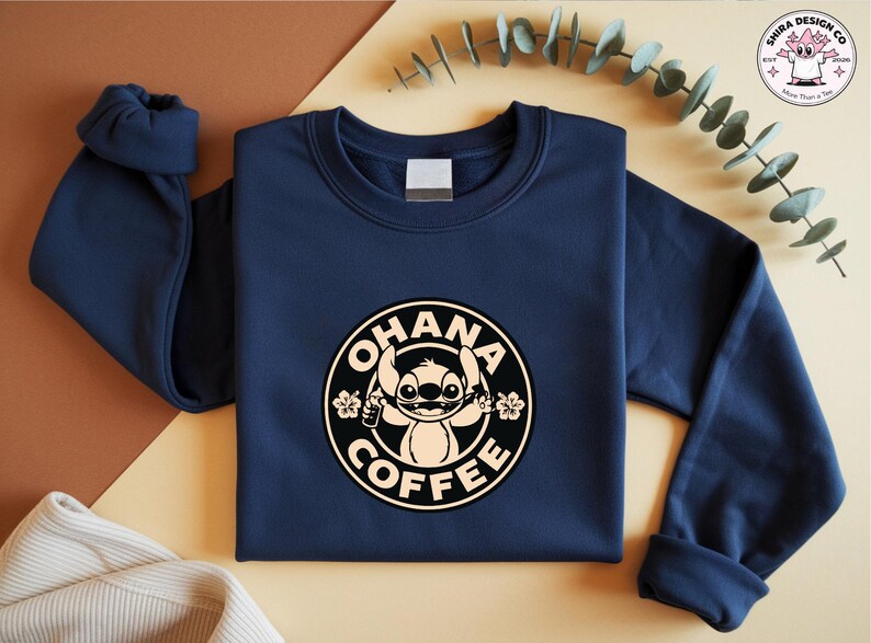 May include: Navy blue sweatshirt featuring a circular graphic with the text "OHANA COFFEE" and a cartoon character. The design is in black and beige. The sweatshirt has long sleeves and a crew neck.