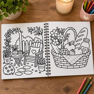 May include: A spiral-bound coloring book open to two pages featuring black and white line art. The left page shows a garden scene with a chair, table, and potted plants. The right page displays a picnic basket filled with fruit, bread, and a bottle.