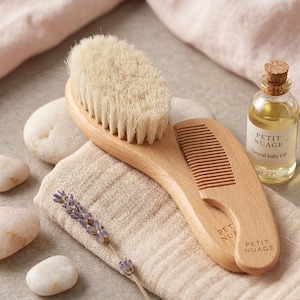 Puede incluir: Un juego de cepillo y peine para bebé de madera con cerdas suaves y un mango curvo. Una pequeña botella de aceite natural para bebé con tapón de corcho está junto al cepillo. El conjunto está sobre una tela beige.