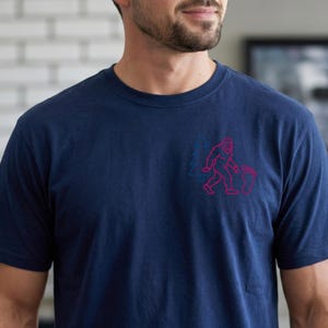 May include: Navy blue t-shirt featuring a Bigfoot graphic in pink and a blue tree design. The Bigfoot is walking with a footprint and a Christmas stocking. The shirt is made of a soft material.