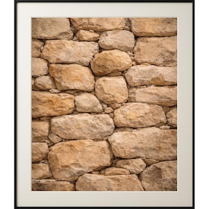 May include: Framed print showing a close-up of a stone wall. The wall is made of various sized, irregularly shaped, beige and tan stones. The stones are tightly fitted together, creating a textured surface. The print is set in a black frame with a white mount.