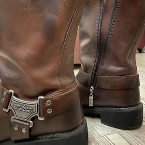 Puede incluir: Un par de botas de cuero marrón a media pantorrilla con suela de goma negra y cremallera lateral. Las botas presentan una placa metálica plateada con el logo de Harley-Davidson en la correa del tobillo y una pequeña etiqueta metálica en el talón.