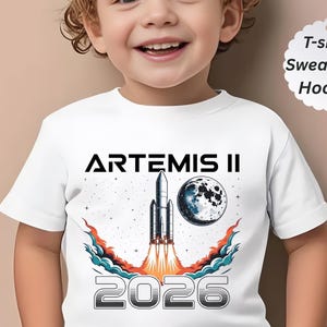 May include: White t-shirt featuring a graphic of a rocket launching into space, with the text "ARTEMIS II" and "2026". The design includes a moon and stylized flames. The shirt is labeled as a t-shirt, sweatshirt, or hoodie.