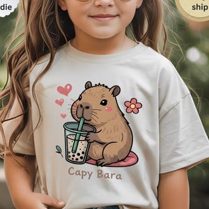 Op de afbeelding: Beige T-shirt met een cartoon capibara die boba thee drinkt. Het ontwerp bevat roze hartjes, een bloem en de tekst "Capy Bara". Het shirt is gelabeld met "T-shirt, Sweatshirt, Hoodie" en "Next day ship".