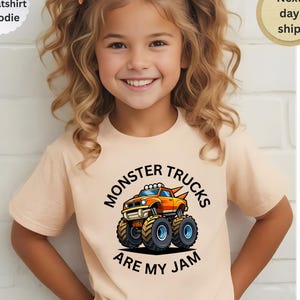 May include: A light peach-colored t-shirt featuring a colorful graphic of a monster truck with the text "MONSTER TRUCKS ARE MY JAM". The shirt is short-sleeved and has a round neckline. The image also includes text that says "T-shirt, Sweatshirt, Hoodie" and "Next day ship".