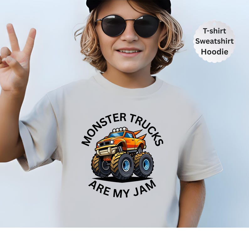 May include: A light beige t-shirt with a colorful graphic of an orange monster truck with blue wheels. The text "MONSTER TRUCKS ARE MY JAM" is printed on the shirt. The shirt is labeled as a t-shirt, sweatshirt, and hoodie.
