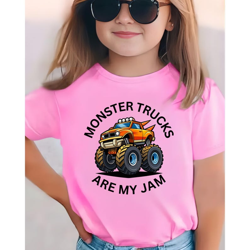 May include: Pink t-shirt featuring a cartoon monster truck in orange and yellow with large black tires. The text "MONSTER TRUCKS ARE MY JAM" is printed in black around the truck. The shirt is worn by a person wearing sunglasses.