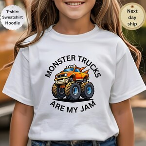 May include: White t-shirt featuring a colorful cartoon monster truck design. The truck is orange and yellow with large wheels, and the text "MONSTER TRUCKS ARE MY JAM" is printed above and below the truck. The shirt is a t-shirt, sweatshirt, or hoodie.
