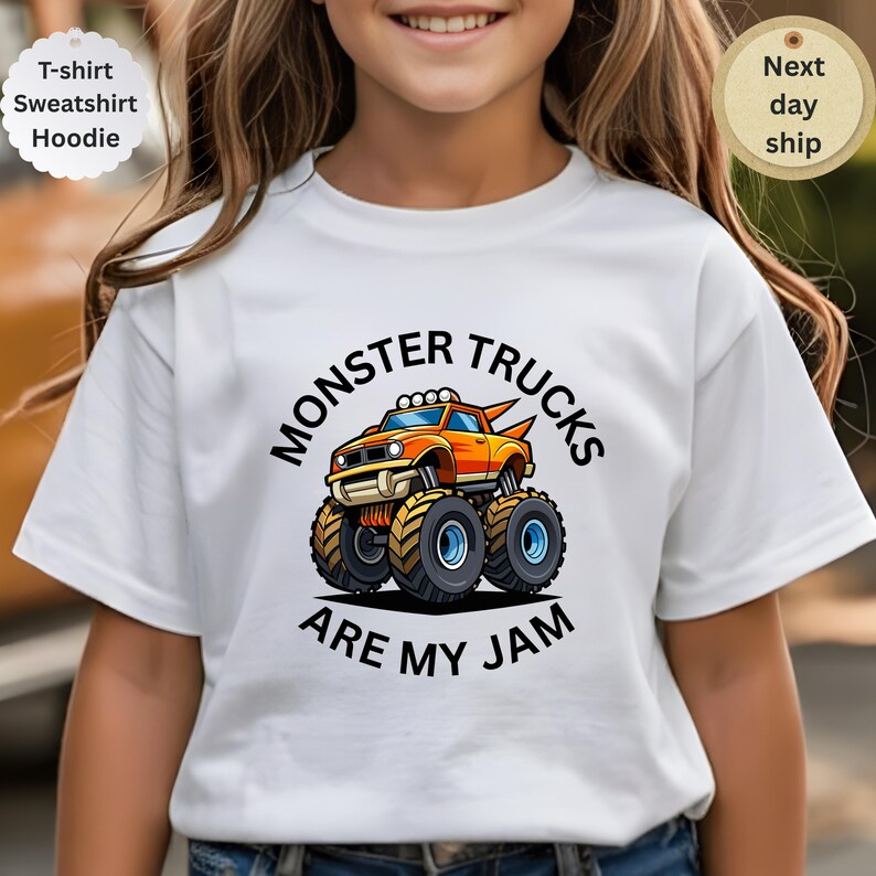 May include: White t-shirt featuring a colorful cartoon monster truck design. The truck is orange and yellow with large wheels, and the text "MONSTER TRUCKS ARE MY JAM" is printed above and below the truck. The shirt is a t-shirt, sweatshirt, or hoodie.